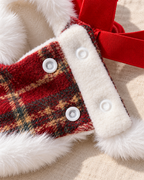 Close-up of a red and beige plaid pet vest with white faux fur trim and white snap buttons, shown on a beige fabric surface.