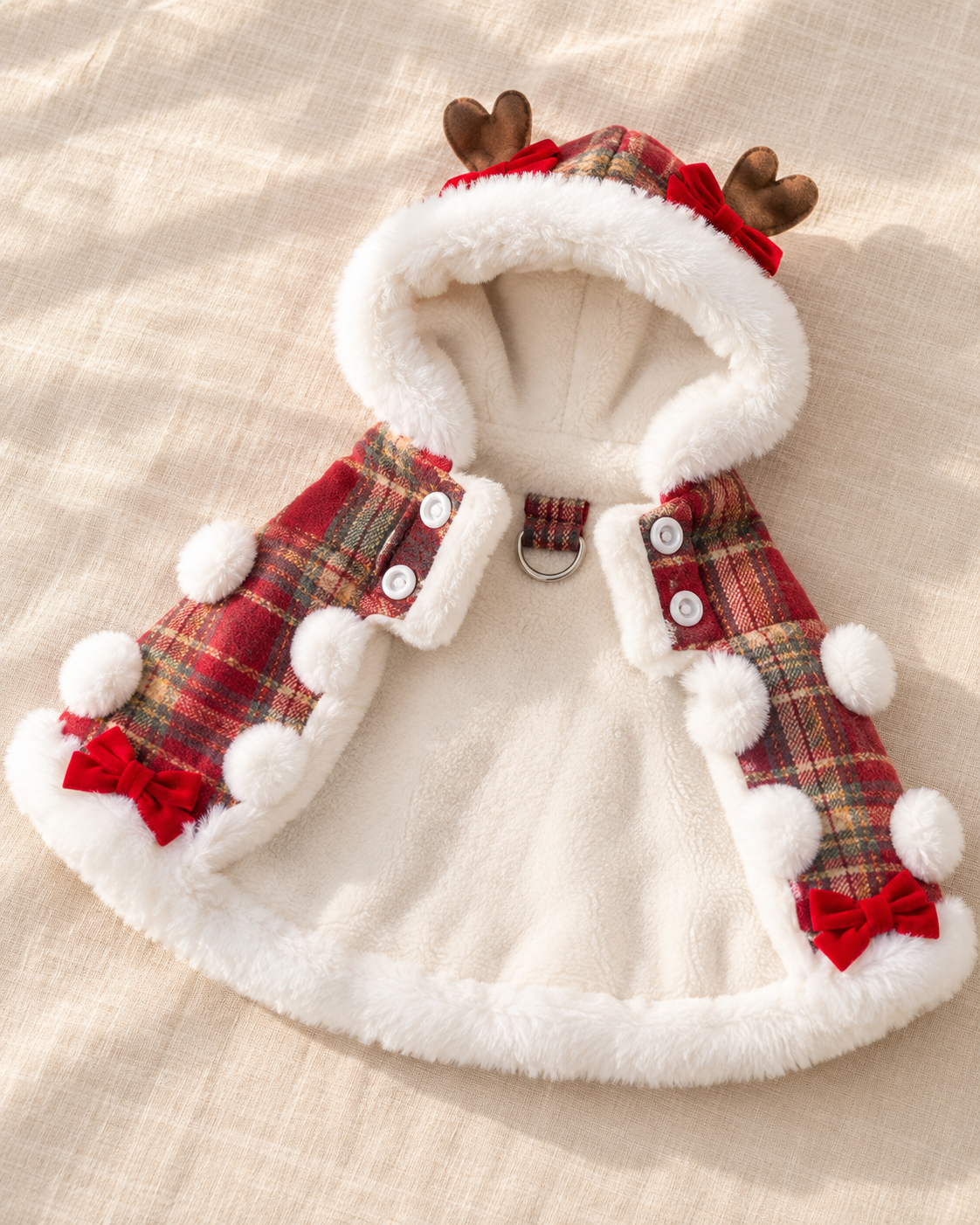 A festive cat's hooded cape or poncho laid flat on a light beige fabric background. The garment has a white faux-fur trim, a red and green plaid pattern on the hood and side panels, white pom-poms, white buttons, reindeer antlers on the hood, and small red bows near the lower sides.