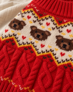 Close-up of a red knitted sweater with a cream-colored yoke featuring brown teddy bear faces, small red hearts, and yellow and dark brown zigzag patterns, plus a red cable-knit lower section.