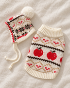 A knitted pet sweater and matching hat laid flat on a cream fabric background. The hat has a fluffy cream pom-pom and drawstrings, while the sweater features a cream base with red apple motifs, red heart patterns, and dark brown geometric bands.